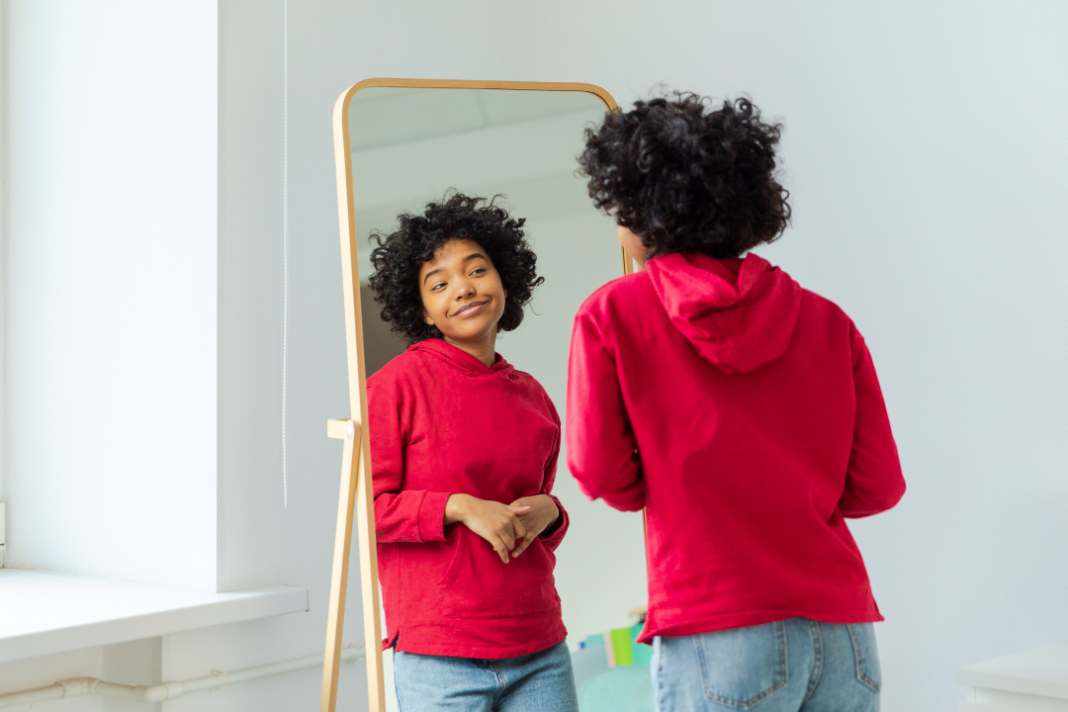Young woman smiling at her reflection in the mirror, representing confidence and the benefits of self-awareness.