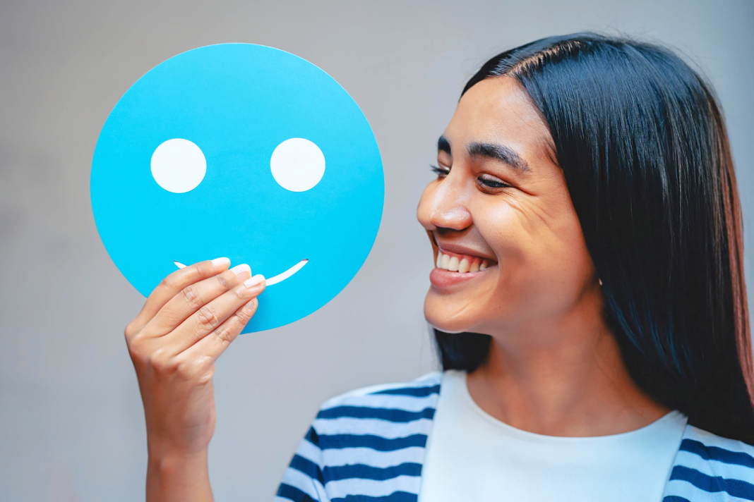 Smiling woman holding a blue happy face cutout, representing confidence and signs of good mental health.